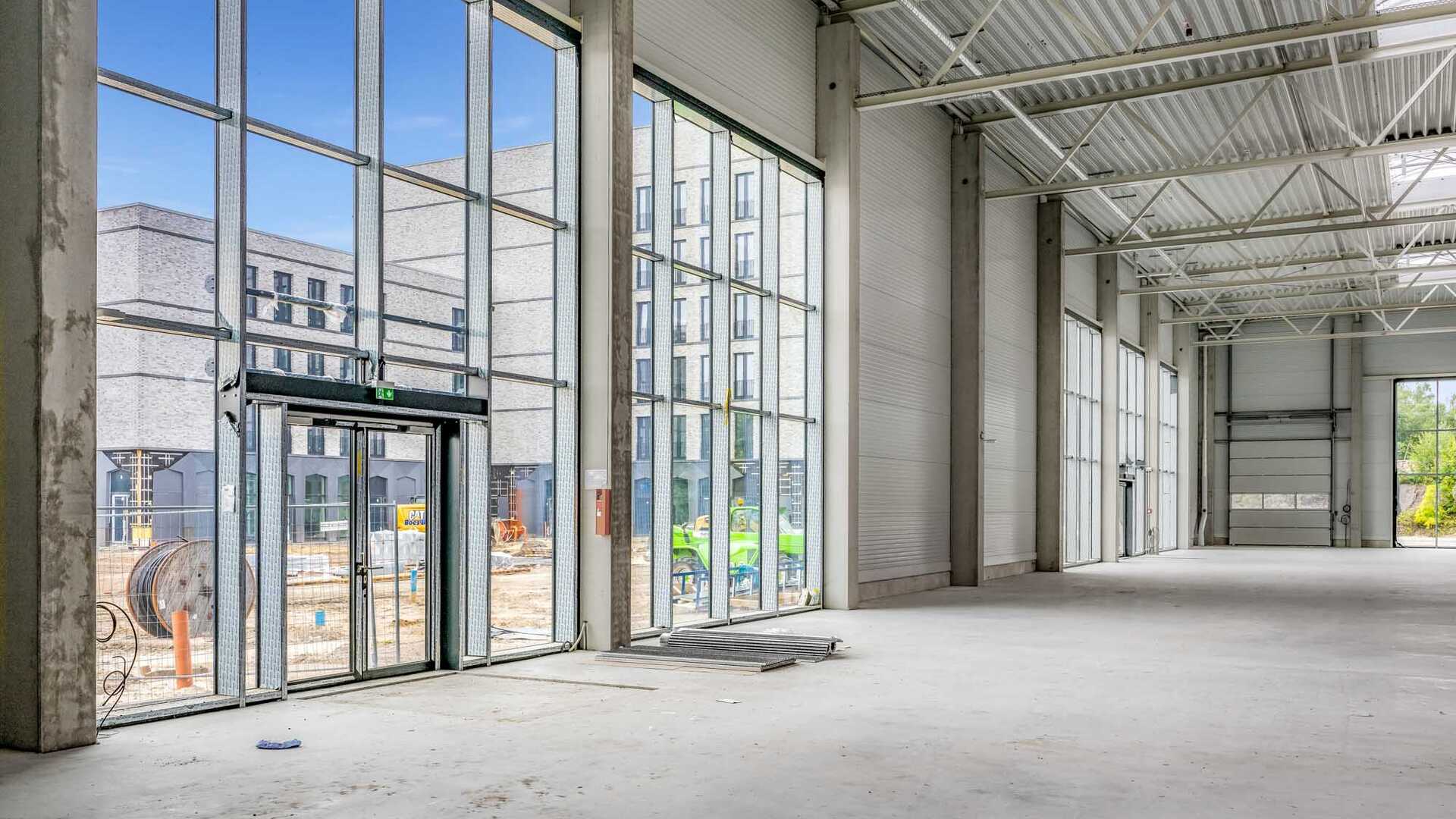 Photo taken during the construction phase of the railway track complex. It shows a large hall with a glass front entrance and high ceilings. Outside the hall, you can see construction equipment and other buildings of the railway yard complex.