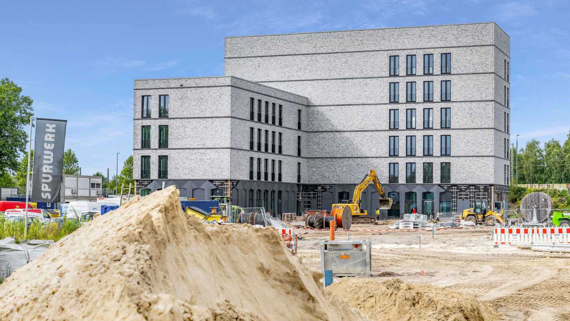 Photograph of the construction process of the trackworks complex. It shows a finished large building made of grey stone, with an excavator in front of it on a large filled-in field.